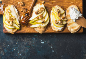 Crostini with pear, ricotta cheese, honey and walnuts. Breakfast toasts or snack sandwiches on rustic wooden board over dark blue grunge plywood background. Top view, copy space